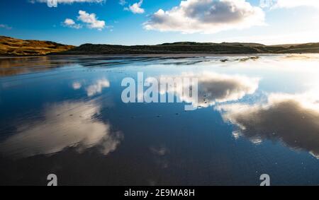 Wolken spiegeln sich an einem nassen Strand an einem sonnigen Tag Stockfoto