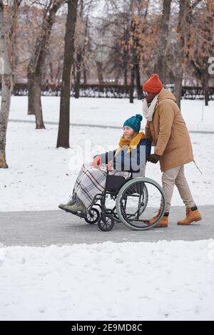 Afrikanischer junger Mann mit behinderter Frau im Rollstuhl, der zusammen läuft Im Freien im Winter Tag Stockfoto