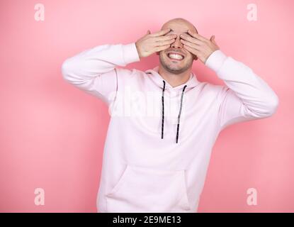 Junge Glatze Mann trägt lässige Sweatshirt über rosa isoliert Hintergrund bedeckt Augen mit Händen lächelnd fröhlich und lustig. Blindes Konzept. Stockfoto