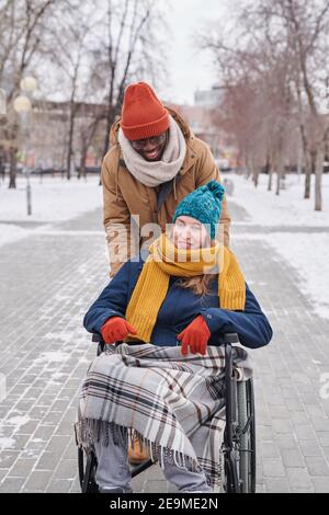 Junge behinderte Frau im Rollstuhl genießen sie zu Fuß auf frisch Luft mit afrikanischem Mann hinter ihr Stockfoto