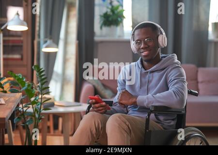 Afrikanischer behinderter Mann in kabellosen Kopfhörern lächelt während der Kamera Musik auf seinem Handy hören Stockfoto