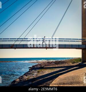 Ein Mann geht an einem sonnigen Wintertag in Fuengirola über eine Hängebrücke vor dem Meer. Stockfoto