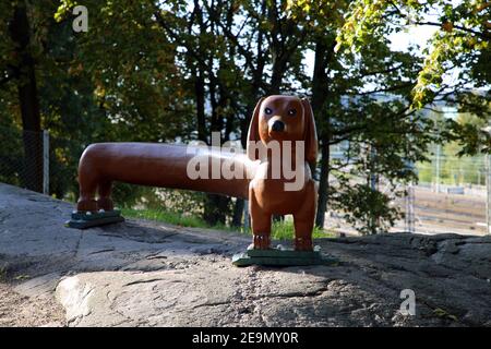 Lange Dackel-förmige braune Bank in einem Hundepark in Helsinki, Finnland. Fotografiert Im September 2019. Die hundeförmige Bank ist auf einer Felswand von Bäumen Stockfoto