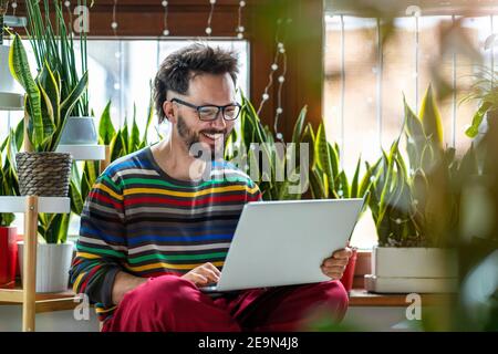 Junger Mann, der zu Hause mit einem Laptop arbeitet und von Zimmerpflanzen umgeben ist Stockfoto