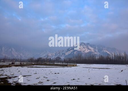 Pendler gehen entlang der Straße mit schneebedeckten Bergen im Hintergrund während eines Winterabends in Srinagar.Wetter über die Stadt Srinagar hat sich verbessert, nachdem Kaschmir frischen Schneefall erhielt. Die Mindesttemperatur blieb über dem Gefrierpunkt in der Stadt Srinagar, da das Wetterbüro das trockene Wetter für die nächsten sieben Tage prognostizierte. Stockfoto