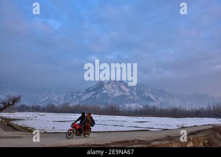 Ein Motorradfahrer fährt entlang der Straße mit schneebedeckten Bergen im Hintergrund während eines Winterabends in Srinagar.Wetter über der Stadt Srinagar hat sich verbessert, nachdem Kaschmir frischen Schneefall erhielt. Die Mindesttemperatur blieb über dem Gefrierpunkt in der Stadt Srinagar, da das Wetterbüro das trockene Wetter für die nächsten sieben Tage prognostizierte. Stockfoto