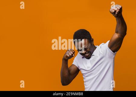 Glücklich schöner schwarzer Mann in weißem Hemd auf orangefarbenem Hintergrund mit leerem Platz links. Gewinnen Sie das Spiel. African dreht seine Hände hoch und schreit laut Stockfoto