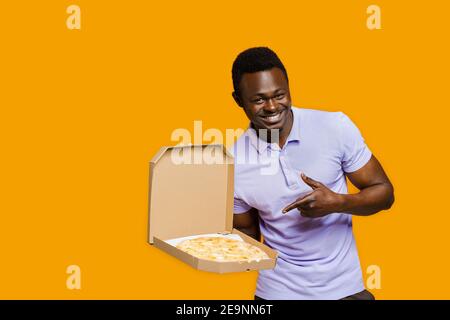 Leckeres Essen Pizzalieferung vom Restaurant. Sicherheitslieferung. Pizza mit Käseplatten werben. Isolierter gelber Hintergrund. Junge afrikanische bärtige Cour Stockfoto