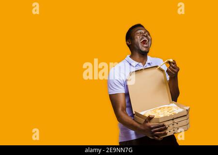 Witziger schwarzer Kurier lachet und hält ein Stück Pizza mit 4 Pizzaboxen. 2+2 großer Rabatt für Kunden. Leckeres Essen Pizzalieferung vom Restaurant. Sa Stockfoto