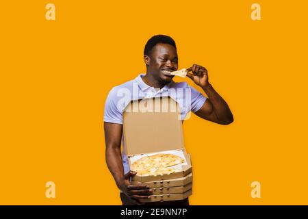 Lustige schwarze Kurier isst Stück Pizza und hält 4 Pizzaboxen. 2+2 großer Rabatt für Kunden. Leckeres Essen Pizzalieferung vom Restaurant. Sicherheitsabsch Stockfoto