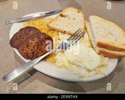 Amerikanisches Frühstück mit Eiern, Kartoffelrösti, Bratwurst und Toast in einem Restaurant. Stockfoto