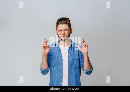 Junger, gutaussehender Mann kreuzt die Finger und macht einen Wunsch. Glücklicher Mann in blauen Jeans-Shirt will in Online-Wette zu gewinnen. Emotionales Porträt kaukasischer Mann isola Stockfoto