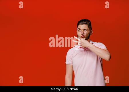 Lustig schöner Mann dumm herum. Denken und verrückte Emotionen machen. Kaukasischer Mann berührt seinen Bart und denkt. Isolierter roter Hintergrund. Online-Arbeit Stockfoto