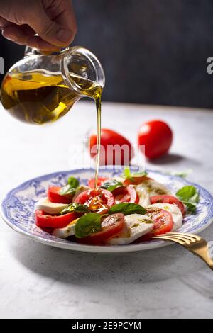 Der Koch gießt Olivenöl auf einen Caprese-Salat. Italienischer berühmter Salat mit frischen Tomaten, Mozzarella-Käse und Basilikum Stockfoto