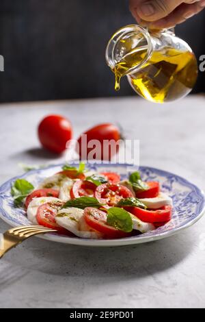Mann gießt Olivenöl auf einen Caprese Salat. Italienischer berühmter Salat mit frischen Tomaten, Mozzarella-Käse und Basilikum Stockfoto