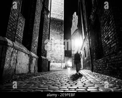 Beleuchtete gepflasterte Straße mit Lichtreflexen auf Kopfsteinpflaster in der alten historischen Stadt bei Nacht. Dunkle verschwommene Silhouette der Person erinnert an Jack the Ripper. Schwarzweiß-Bild. Stockfoto