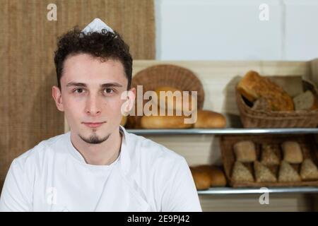 Hinter der Theke seines Ladens steht ein großer und hübscher Bäcker in einem Bäckeroutfit. Familienbäckerei mit mehrgenerationenübergreifenden Traditionen. Stockfoto