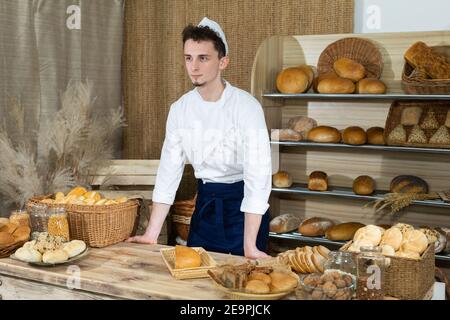 Hinter der Theke seines Ladens steht ein großer und hübscher Bäcker in einem Bäckeroutfit. Familienbäckerei mit mehrgenerationenübergreifenden Traditionen. Stockfoto