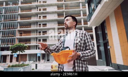 Junge kaukasische männliche Manager und Supervisor auf der Baustelle leitet Prozess und gibt Anweisungen an untergeordnete Arbeitnehmer. Leitender Ingenieur gibt Stockfoto