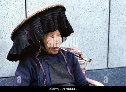 Hongkong, China. Eine Hakka-Frau, die vor einem Tempel in den Neuen Territorien eine Pfeife raucht und einen traditionellen Hut trägt. Stockfoto