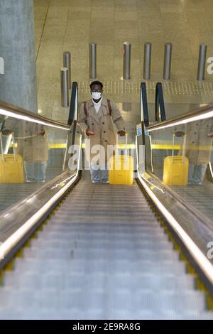 Schwarzer Mann mit Koffer steht auf Rolltreppe im Flughafen tragen Gesichtsmaske während covid-19 Pandemie. Stockfoto