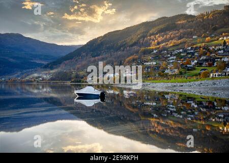 Ein Boot schwimmt in einem See in Voss, Norwegen, das stille Wasser sieht aus wie ein Spiegel, spiegelt das Haus auf dem Hügel mit den Bäumen im Herbst am Vorabend Stockfoto