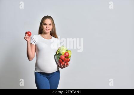 Schwangere blonde Frau hält frisches Gemüse in einer Schüssel und Tomate in der Hand Stockfoto