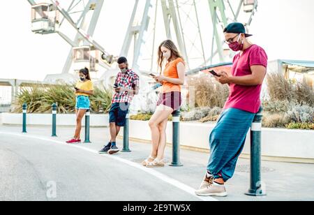 Multiethnische Freunde tragen Gesichtsmaske mit Tracking-App mit Handy Smartphone - gelangweilte junge Menschen am Riesenrad - Neuer normaler Lebensstil Stockfoto