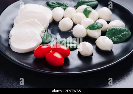 Weiße kleine Mozzarella Käsebällchen, Spinatblätter und Tomaten auf schwarzem Teller. Stockfoto