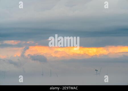 Windturbinen des Offshore-Windparks London Array, die aus der Morgendämmerung hervorgehen, Nebel am frühen Morgen vor der Küste von Kent in Herne Bay. Die Spitzen der Turbinen sind mit einem bewölkten, grauen Himmel mit orangefarbenen Streifen sichtbar. Das Meer ist ruhig mit Nebel in einer Schicht darüber. Stockfoto
