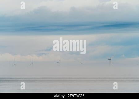 Windturbinen des Offshore-Windparks London Array, die aus der Morgendämmerung hervorgehen, Nebel am frühen Morgen vor der Küste von Kent in Herne Bay. Die Spitzen der Turbinen sind mit einem wolkigen, grauen Himmel sichtbar. Das Meer ist ruhig mit Nebel in einer Schicht darüber. Stockfoto