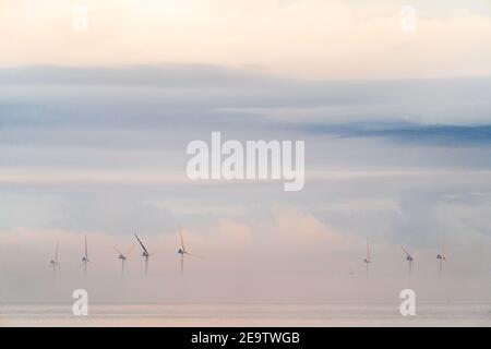 Windturbinen des Offshore-Windparks London Array, die aus der Morgendämmerung hervorgehen, Nebel am frühen Morgen vor der Küste von Kent in Herne Bay. Die Spitzen der Turbinen sind mit einem wolkigen, grauen Himmel sichtbar. Das Meer ist ruhig mit Nebel in einer Schicht darüber. Stockfoto