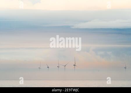 Windturbinen des Offshore-Windparks London Array, die aus der Morgendämmerung hervorgehen, Nebel am frühen Morgen vor der Küste von Kent in Herne Bay. Die Spitzen der Turbinen sind mit einem bewölkten, grauen Himmel mit orangefarbenen Streifen sichtbar. Das Meer ist ruhig mit Nebel in einer Schicht darüber. Stockfoto