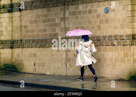 Frau mit rosa Regenschirm und tragen Leggings und modischen Mantel geht auf dem Bürgersteig, wie sie aus den Geschäften im Regen zurückkehrt. Stockfoto