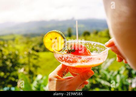 Detail eines Cocktailglases, das von einer Frau vor einer Waldlandschaft getrunken wird. Costa Rica Gastronomie Stockfoto