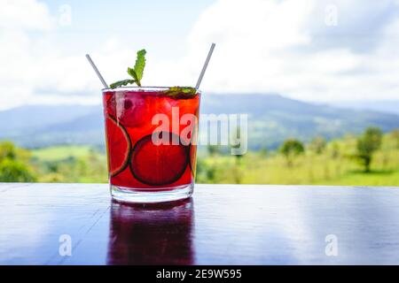 Cocktailglas mit roten Früchten, verziert mit Zitrone und Minze Ein Holztisch mit einem Wald im Hintergrund Stockfoto