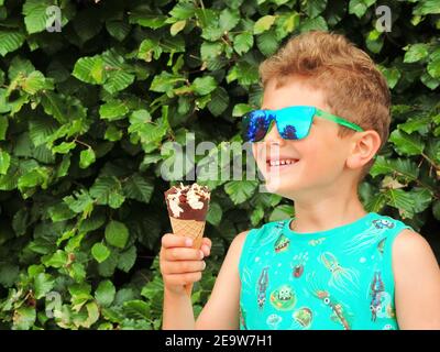 Liebenswert kleiner Junge in Sonnenbrille hält ein Eis-Kegel In seinen Händen und lächelt Stockfoto