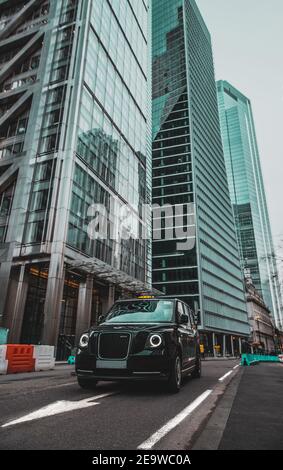 London UK Januar 2021 Vertikale Aufnahme eines modernen Hybrid-londoner Taxis, das an der leadenhall Street vorbeifährt, riesige Hochhäuser, die hoch in der Stadt emporragen Stockfoto