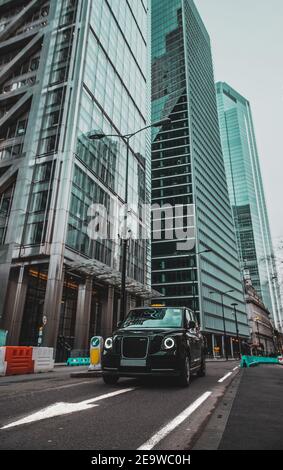 London UK Januar 2021 Vertikale Aufnahme eines modernen Hybrid-londoner Taxis, das an der leadenhall Street vorbeifährt, riesige Hochhäuser, die hoch in der Stadt emporragen Stockfoto