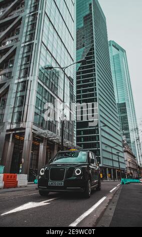 London UK Januar 2021 Vertikale Aufnahme eines modernen Hybrid-londoner Taxis, das an der leadenhall Street vorbeifährt, riesige Hochhäuser, die hoch in der Stadt emporragen Stockfoto