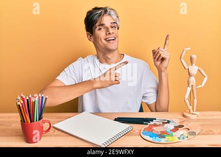 Der junge hispanische Künstler sitzt lächelnd im Studio und schaut auf die Kamera, die mit zwei Händen und Fingern zur Seite zeigt. Stockfoto