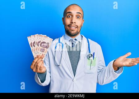 Hispanic erwachsenen Arzt Mann trägt medizinische Uniform hält 500 mexikanische Pesos feiern Leistung mit glücklichen Lächeln und Sieger Ausdruck mit Erhöht Stockfoto