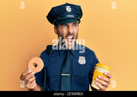 Gutaussehende hispanische Polizei Mann essen Donut und trinken Kaffee wütend und wütend schreien frustriert und wütend, schreien vor Wut. Wut und aggressiv Stockfoto