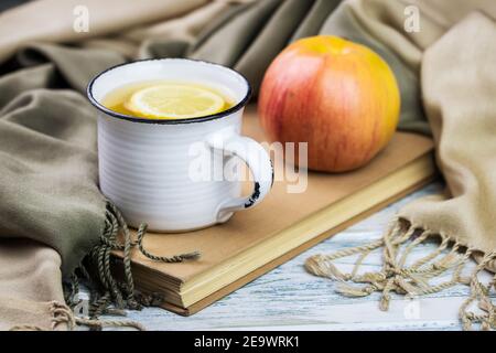 Teetasse und Apfel auf Buchhülle mit Schal, Stillleben auf weißem Holztisch. Frisches Bio-Obst und Heißgetränk Erfrischung. Stockfoto