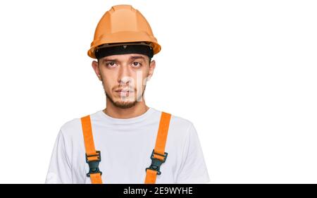 Hispanic junger Mann trägt Handyman Uniform und Sicherheit Hardhut mit ernsten Ausdruck auf Gesicht. Einfach und natürlich Blick auf die Kamera. Stockfoto