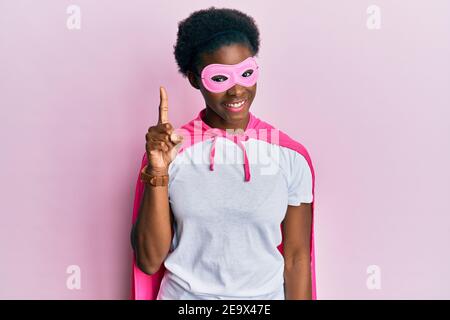 Junge afroamerikanische Mädchen trägt Superhelden Maske und Cape Kostüm zeigt und zeigt mit Finger Nummer eins, während lächelnd zuversichtlich und glücklich. Stockfoto