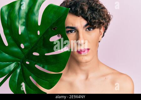 Junger Mann trägt Frau Make-up hält grüne Pflanze Blatt in der Nähe von schönen Gesicht skeptisch und nervös, runzelte wegen des Problems. Negative Pers Stockfoto