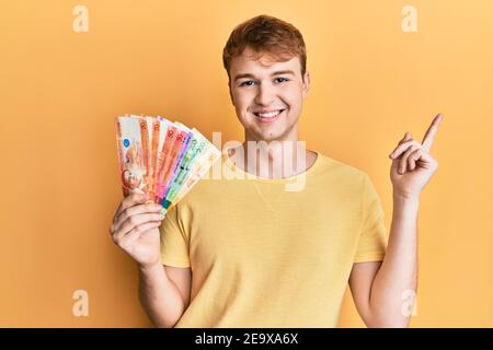 Junger Kaukasianer hält philippinische Peso-Banknoten lächelnd glücklich zeigt Mit Hand und Finger zur Seite Stockfoto