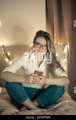 Flacher Fokus einer erwachsenen Frau mit Brille auf der sitzt Ein Bett und SMS an ihr Telefon Stockfoto