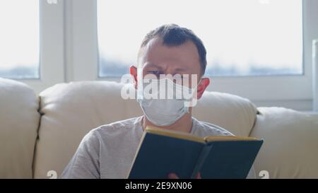 Attraktiver Mann in Maske mit einem Buch in den Händen Schaut auf die Kamera, während sie auf einem Sofa sitzt Stockfoto